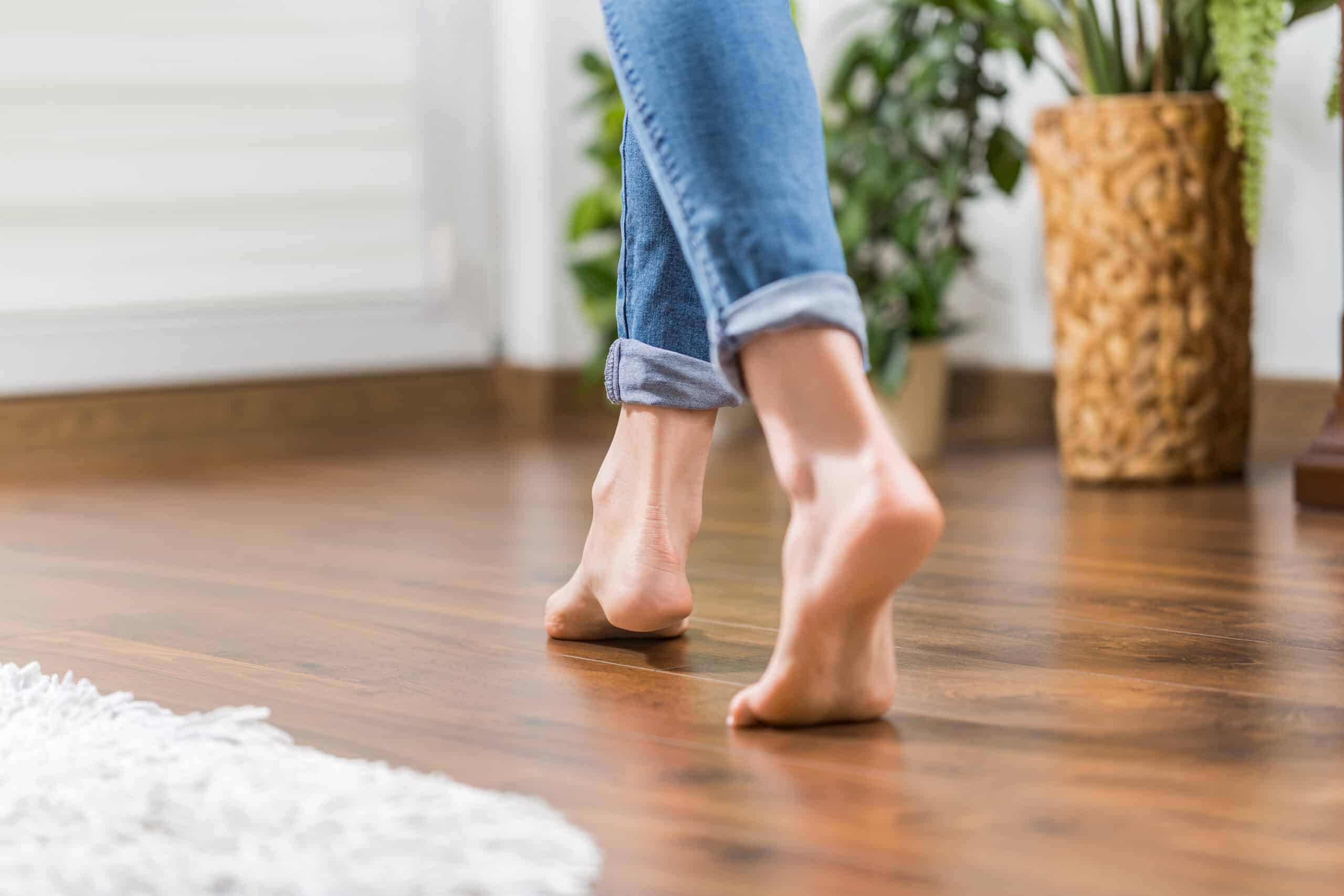 Pieds nus marchant sur un parquet brun chaud, jambes vêtues d'un jean retroussé près d'un tapis blanc.